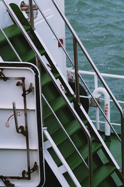 Close-up view of a vessel's green metal staircase with black anti-slip treads, metal handrails on both sides, and white structural supports, leading up from the deck area which contains a white hatch with mechanical fittings. The background shows the water surface, indicating the vessel is stationary or moving on a body of water. The surrounding environment and lighting suggest daytime conditions. This image, associated with home relocation and furniture transport services by Man with Van Aldborough Hatch, highlights the loading process often involved in moves that require shipping or transport via boat as part of the moving logistics.