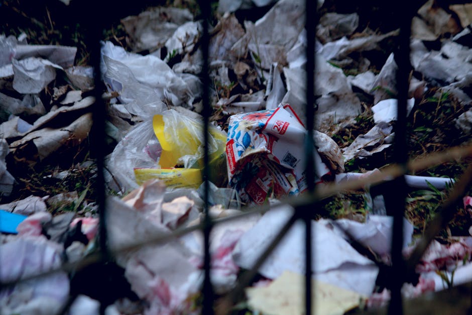 Photograph showing a pile of household waste and discarded packaging materials, including crumpled paper, plastic bags, cardboard boxes, and torn packaging, located on the ground behind a black metal fence. The waste appears to be a mixture of food wrappers, paper, and plastic debris, with some items partially buried in dried leaves and grass. The environment suggests an outdoor area near a property or driveway, with natural lighting. This image highlights improper disposal of waste, which [COMPANY_NAME] in Aldborough Hatch can assist with during home relocation or furniture transport by providing proper waste management and removal services, ensuring clean and compliant disposal of household items during packing and moving processes.