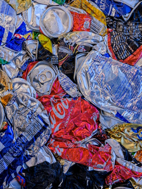 A close-up view of a pile of crushed and flattened aluminum beverage cans, including soda cans with visible pull-tabs and colorful branding, mixed with some plastic and paper packaging materials. The cans are irregularly shaped, overlapping each other, and reside on a surface likely within a recycling or waste collection area. The scene highlights materials typically associated with waste management and rubbish disposal, relevant to house removals or relocation services provided by Man with Van Aldborough Hatch. This image exemplifies waste sorting and disposal processes involved in home relocation or clearance tasks, emphasizing the importance of proper waste separation before moving or disposal in the Aldborough Hatch area.