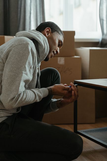 A man with short dark hair and medium skin tone is kneeling on the floor of a room during a home relocation process, examining his hands with a focused expression. He is wearing a light grey hoodie and black pants. Behind him, there are several cardboard moving boxes of various sizes, some sealed with tape, arranged near a large window with natural light streaming in, illuminating the scene. The boxes appear to be part of packing and moving preparations, inside a residential space possibly in the process of unpacking or furniture assembly after a relocation. A dark-colored table is visible adjacent to him, with part of it in the frame. In the context of furniture assembly and moving services offered by Man with Van Aldborough Hatch, this scene highlights the post-move phase involving unpacking, organizing, or fixing furniture and household items as part of a professional house removal service.