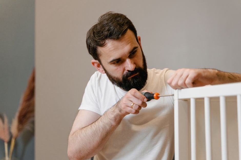 A man with a beard and dark hair, wearing a white t-shirt, is using a screwdriver to assemble or fix a white wooden crib indoors. The man is focused on the task, holding the screwdriver with his right hand while stabilising the crib with his left hand. The cribs are located on a light-colored floor beside a plain, neutral-toned wall. In the background, there are packaging materials such as cardboard boxes and plastic wrapping, hinting at ongoing packing or furniture assembly during a house move. The lighting appears to be natural or from indoor fixtures, illuminating the workspace clearly. This image relates to furniture assembly as part of house removals or moving services, illustrating the process of reassembling furniture after transportation, which Man with Van Aldborough Hatch provides as part of their relocation solutions.