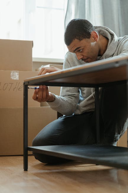 A man with short dark hair and medium skin tone is kneeling on the floor of a room during a home relocation process, examining his hands with a focused expression. He is wearing a light grey hoodie and black pants. Behind him, there are several cardboard moving boxes of various sizes, some sealed with tape, arranged near a large window with natural light streaming in, illuminating the scene. The boxes appear to be part of packing and moving preparations, inside a residential space possibly in the process of unpacking or furniture assembly after a relocation. A dark-colored table is visible adjacent to him, with part of it in the frame. In the context of furniture assembly and moving services offered by Man with Van Aldborough Hatch, this scene highlights the post-move phase involving unpacking, organizing, or fixing furniture and household items as part of a professional house removal service.