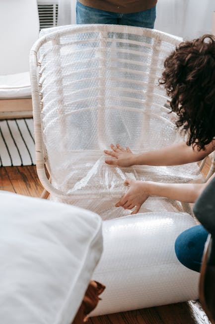 A person is assembling a white wicker laundry basket inside a room, with a focus on their hands positioning a clear plastic lining into the basket. The room features wooden flooring, and part of a bed with white bedding and a brown blanket is visible in the foreground. In the background, a portion of a person wearing blue jeans and an indoor setting with natural lighting can be seen. This scene depicts the process of preparing household items for packing and moving, typical of home relocation and furniture transport services provided by Man with Van Aldborough Hatch. The image emphasizes careful packing of household essentials as part of a professional removals or moving service.
