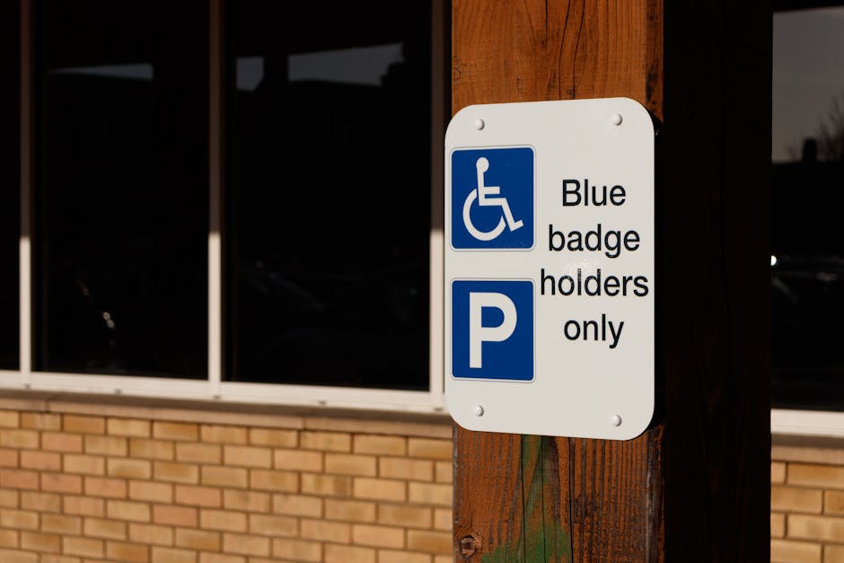 A white rectangular parking sign mounted on a dark brown wooden post outside a building with large glass windows and a brick facade. The sign features a blue wheelchair symbol at the top, followed by the text 'Blue badge holders only', and a large blue 'P' symbol at the bottom. The sign indicates designated parking accessible for disabled individuals with blue badges. The surrounding environment is well-lit, and the signage is clearly visible for accessibility and parking regulations relevant to house removals or relocation services, as handled by Man with Van Aldborough Hatch.