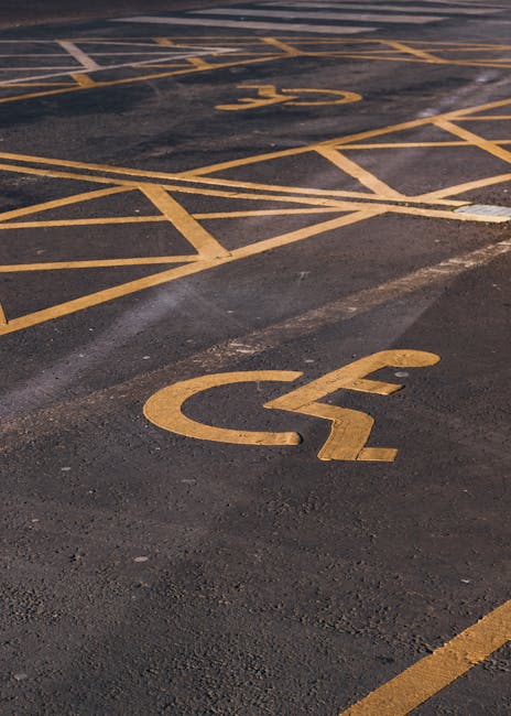 A close-up view of a parking lot surface showing two designated disabled parking bays marked with yellow paint, each featuring a wheelchair symbol. The markings are positioned on dark asphalt, with the lower bay in sharp focus and the upper bay slightly blurred. The parking bays are outlined by parallel yellow lines, with additional diagonal lines creating a structured layout for accessible parking spaces. The surface appears clean with minor tire marks, and the lighting suggests an outdoor setting during daytime. This image is relevant to house removals and moving services by illustrating accessible parking options for loading and unloading furniture and boxes during home relocation, as provided by Man with Van Aldborough Hatch.