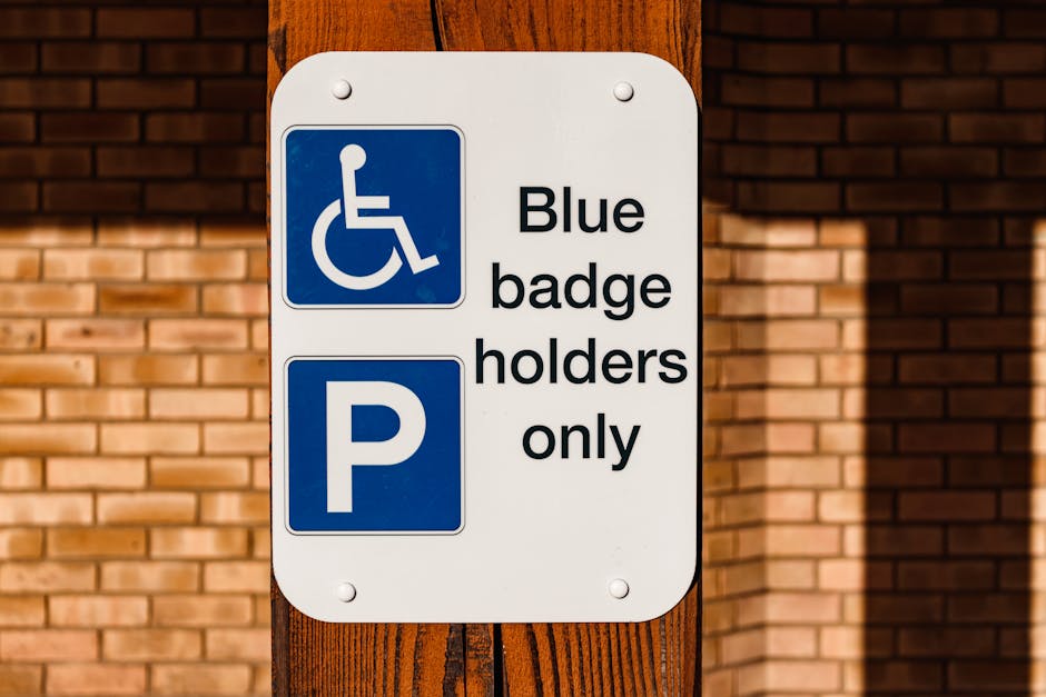 A white rectangular parking sign mounted on a dark brown wooden post outside a building with large glass windows and a brick facade. The sign features a blue wheelchair symbol at the top, followed by the text 'Blue badge holders only', and a large blue 'P' symbol at the bottom. The sign indicates designated parking accessible for disabled individuals with blue badges. The surrounding environment is well-lit, and the signage is clearly visible for accessibility and parking regulations relevant to house removals or relocation services, as handled by Man with Van Aldborough Hatch.