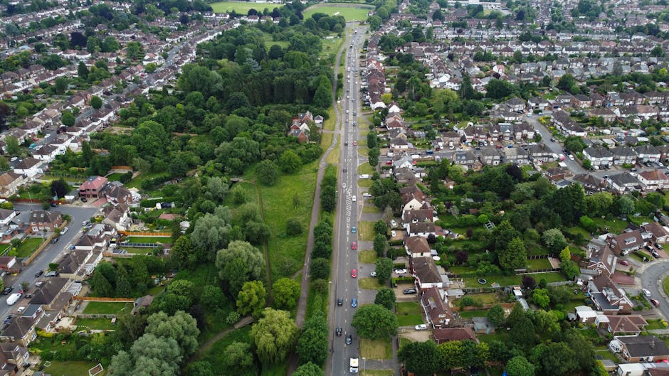 An aerial view of a residential urban area showing a busy main road running through the centre, with multiple lanes of traffic including cars moving in both directions. On one side of the road, there are rows of closely spaced terraced houses with small front gardens, each featuring driveways, some with parked vehicles, and various trees and shrubs. On the opposite side, there are detached and semi-detached houses with larger gardens and greenery, including mature trees, bushes, and lawned areas. The scene includes a strip of green parkland with dense trees and open grass field, positioned adjacent to the road, providing a natural boundary. The lighting is bright, suggesting daytime with clear weather conditions, and the overall environment appears quiet despite the traffic. This image visualises the typical urban streetscape involved in house removals, highlighting property types, packing and moving logistics, and transportation elements, as may be relevant to the services provided by Man with Van Aldborough Hatch for relocating homes within the area.