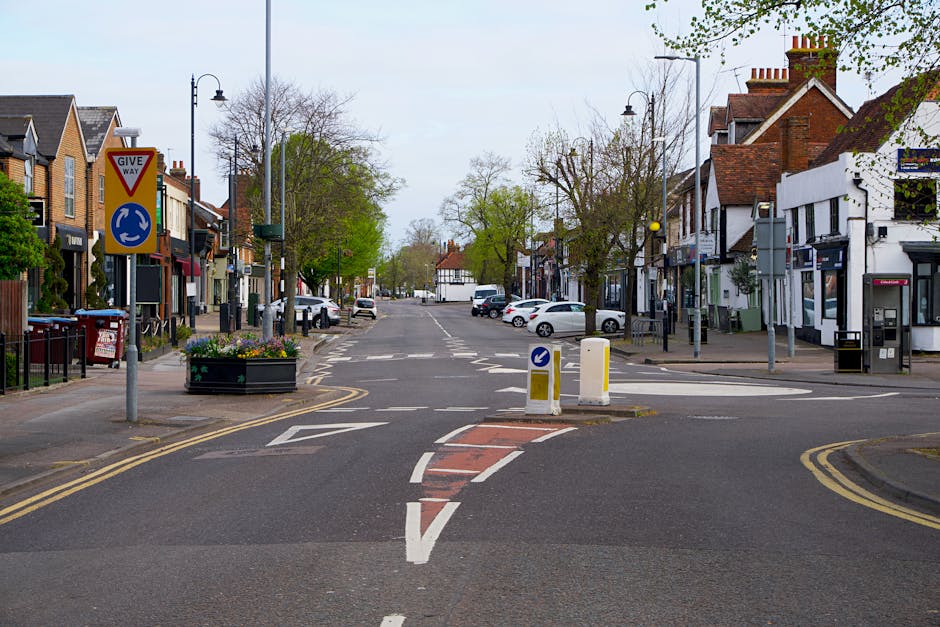 A quiet residential street in Aldborough Hatch with a central roundabout, flanked by shops and houses on both sides. The street is lined with parked cars, and there are traffic signage indicating the roundabout and give way priority. Trees with sparse foliage are visible along the sidewalks, and the sky is overcast, providing diffuse natural lighting. The roadway features marked lanes, a pedestrian crossing, and temporary traffic bollards at the roundabout entrance, indicating potential for traffic management during home relocation or furniture transport activities. Some shop fronts have large windows and outdoor fixtures, while the pavement is wide enough for loading and unloading. This scene illustrates the typical environment where house removals and packing and moving operations by [COMPANY_NAME] such as loading furniture and boxes into a van could take place, especially in areas where careful navigation and adherence to local traffic rules are necessary during moving logistics.