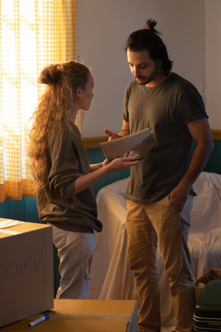 A young woman and a man with long dark hair and beard are engaged in a conversation inside a room with natural light coming through yellow gingham curtains. The woman, with long curly hair tied back, is holding a brown cardboard box, and they are discussing its contents. Behind them, there is a bed with white bedding, and a teal headboard. To the left, a cardboard box with the word 'kitchen' written on it is visible on a wooden table, along with a marker. The man is casually dressed in a grey t-shirt and beige trousers, with his hand in his pocket, while the woman wears a loose jacket and light-colored pants. The scene appears to be part of a home relocation or packing process, with the pair preparing items for moving, supported by the presence of packing materials and unpacked boxes, indicative of a furniture transport and moving logistics context as handled by professionals like Man with Van Aldborough Hatch.