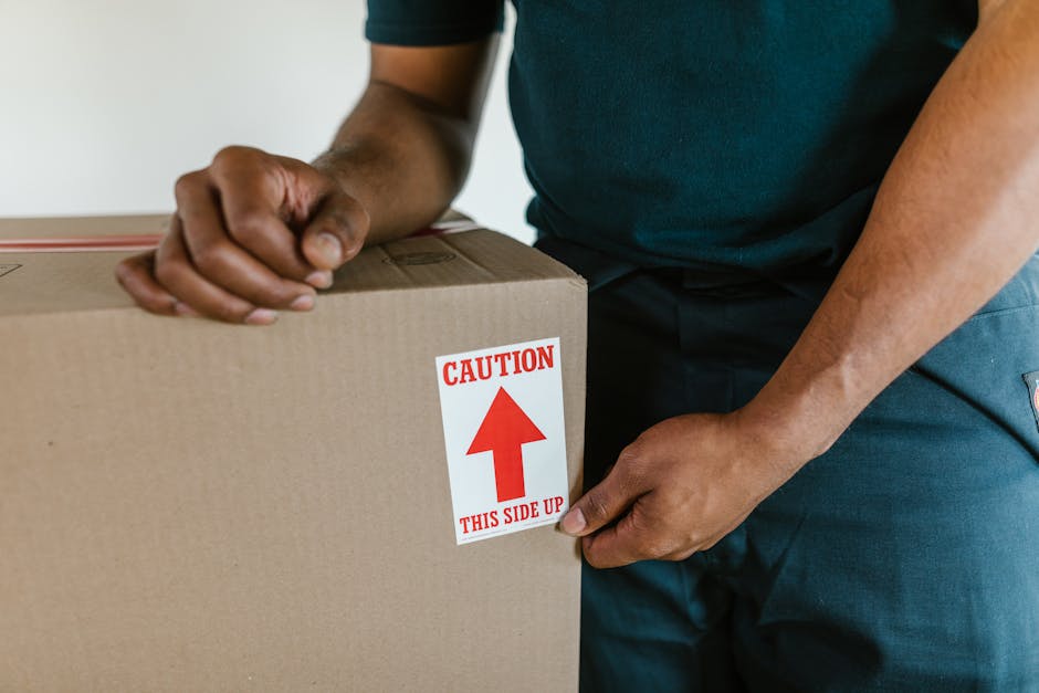 Close-up of a person wearing a dark blue T-shirt and matching trousers during a home relocation, holding a plain cardboard box with a red and white safety label that reads 'Caution This Side Up' with an upward-pointing arrow. The individual's hands are grasping the box securely, with the background showing an indoor environment and possibly a doorway or corridor, indicating the loading or unloading process. The cardboard box appears sturdy, wrapped in plastic, and is part of the packing materials used for furniture transport and packing during a house move. This scene illustrates the careful handling of goods by a professional removals service, such as Man with Van Aldborough Hatch, involved in a packing and moving operation, emphasizing the importance of proper marking for safe transport and proper loading procedures in the relocation process.
