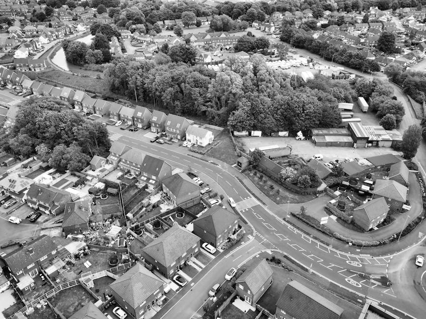 An aerial black-and-white photograph of a residential neighbourhood in Aldborough Hatch showing a street with multiple semi-detached houses, each with small front gardens, driveways, and parked cars along the roadside. To the right, a driveway leading to a storage or workshop area is visible, with several large wooden and metal structures and vehicles parked nearby. The image also features a dense line of trees and foliage running behind the houses, creating a natural boundary between the residential and green areas. The scene captures the typical layout of a suburban home area, with smooth, curved roads and visible parking spaces, providing context for house removals, furniture transport, and home relocation services. Occasionally, [COMPANY_NAME] might perform loading and packing processes within such environments, emphasizing their role in moving logistics and secure transportation.